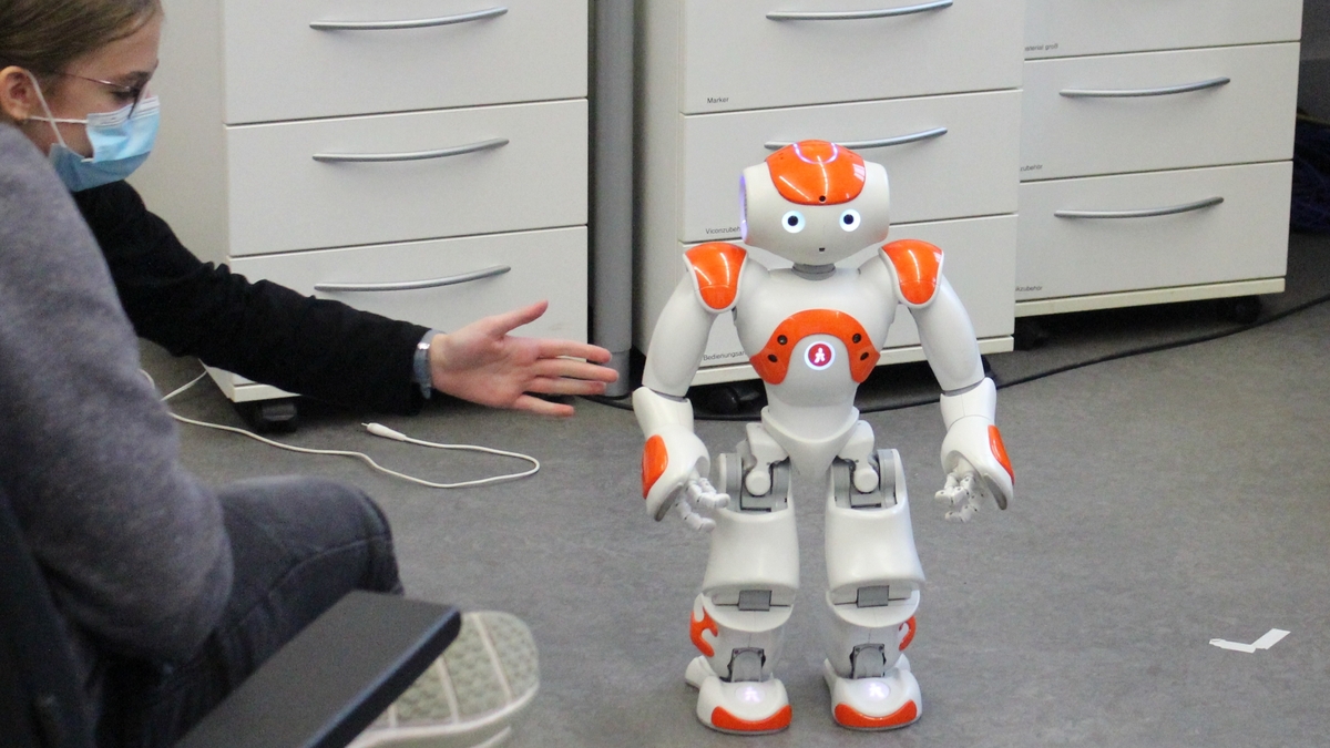 Ein NAO Roboter steht im Zentrum des Bildes, ein Mädchen streckt ihre Hand aus, damit er nicht umfällt